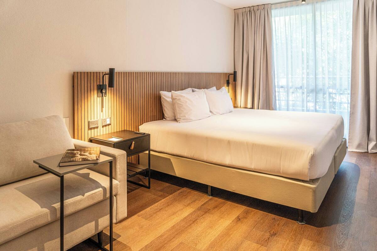 Una habitación de hotel con cama, mesita de noche, mesa, sofá y ventana con cortinas.