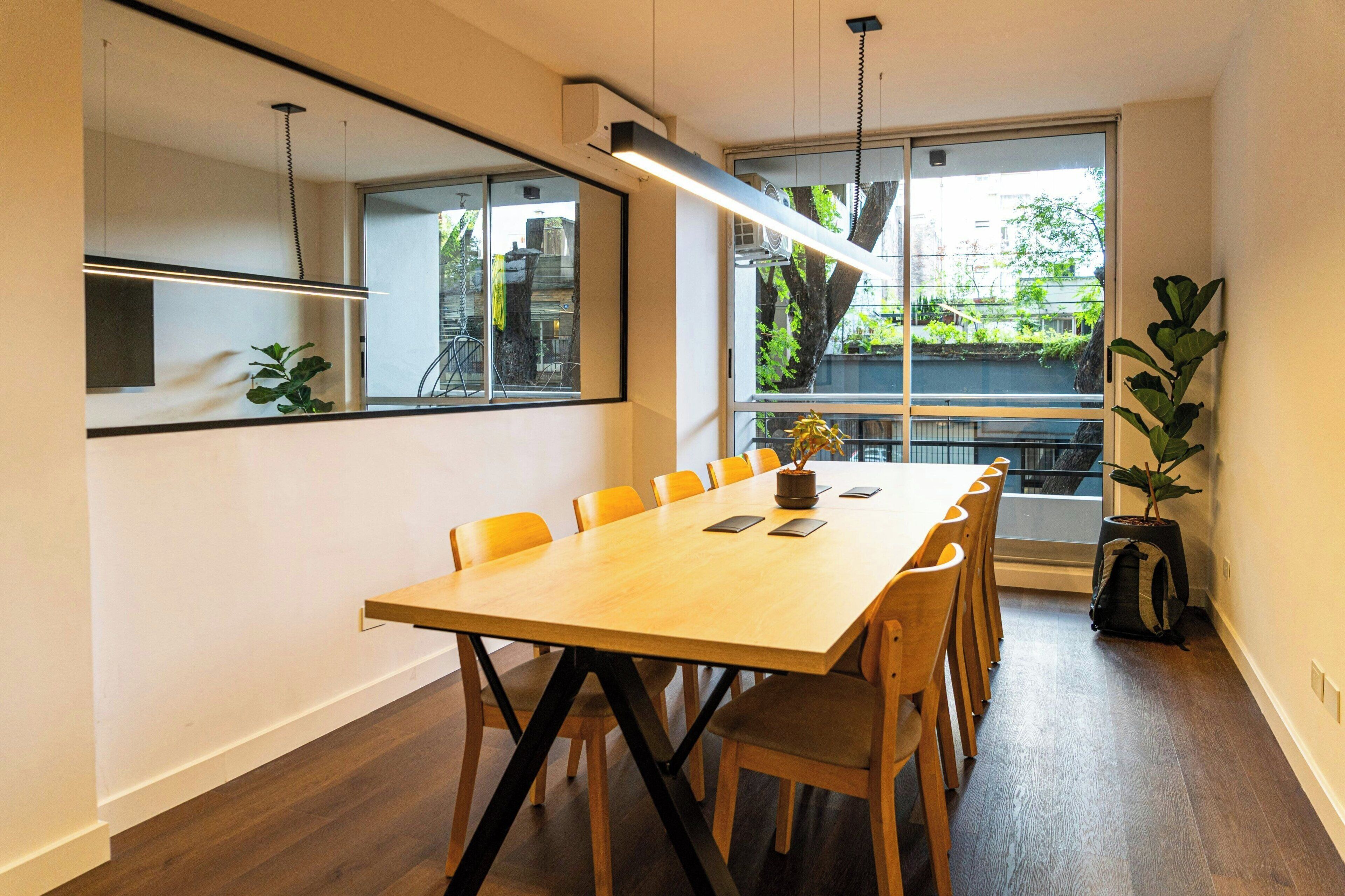 Uma sala de conferências com uma mesa comprida, cadeiras, um vaso de planta e vista através de grandes janelas.