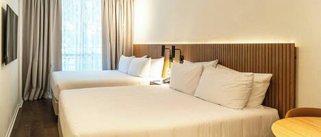 A hotel room with two beds, a wooden headboard, a bedside table, a chair, and a large window with curtains.
