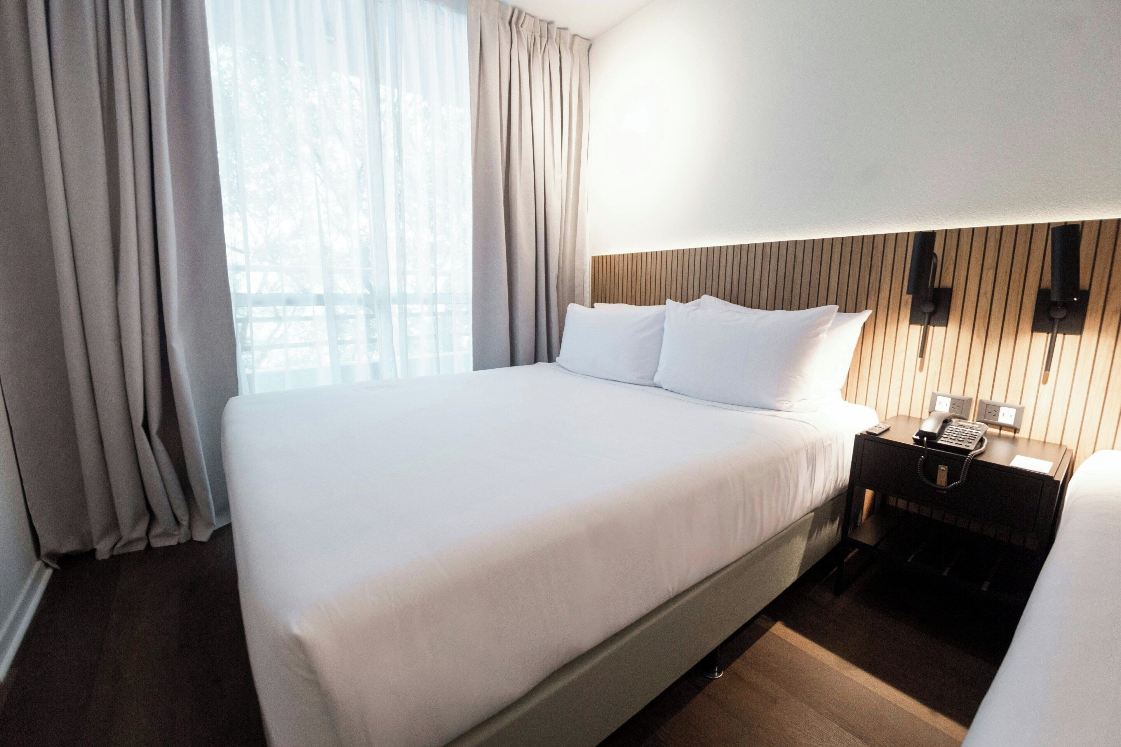 Habitación de hotel con una cama grande, una mesita de noche con teléfono y lámpara, y una ventana con cortinas.