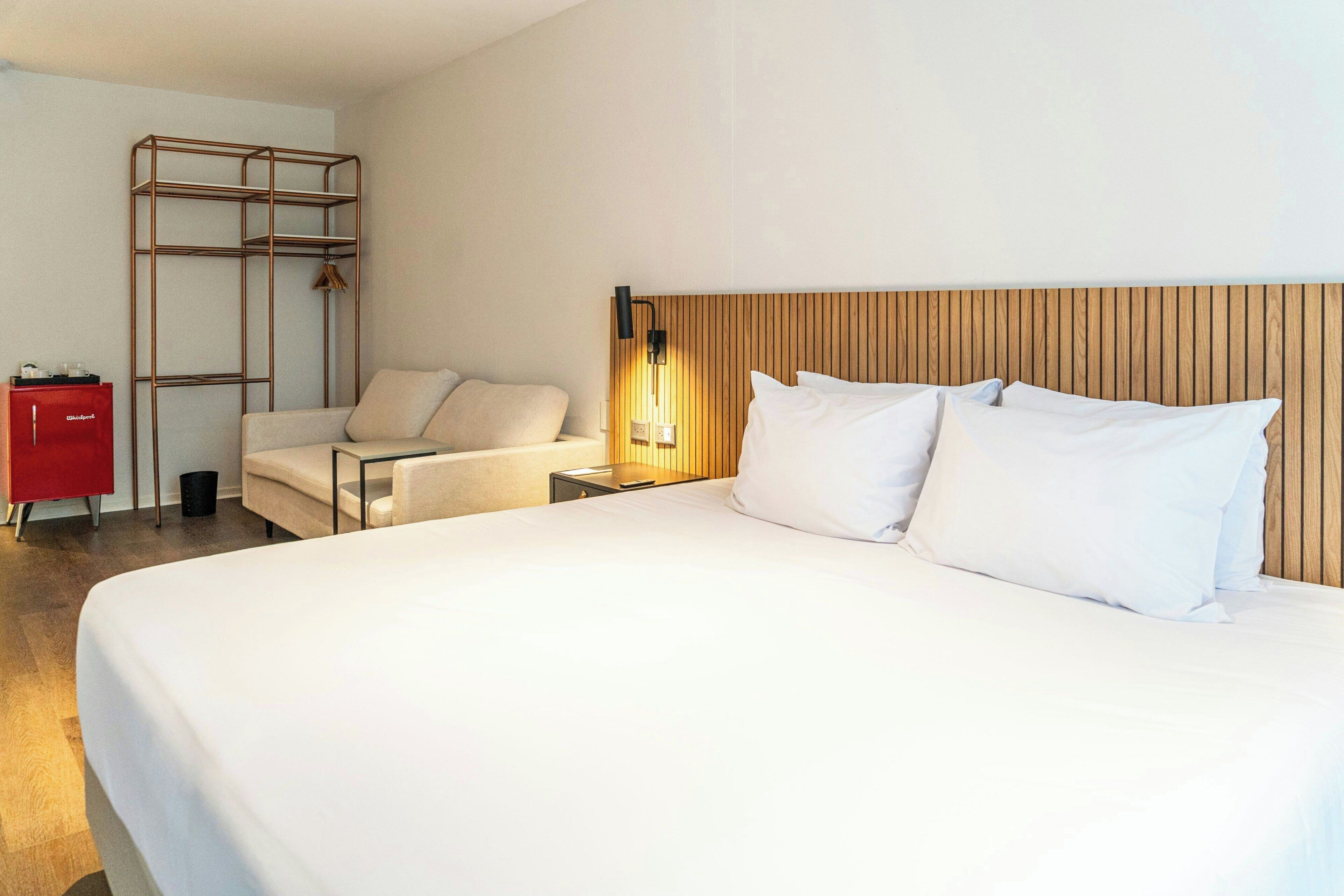 A hotel room with a large bed, a wooden headboard, a red cabinet, a chair, and a metal shelf unit.
