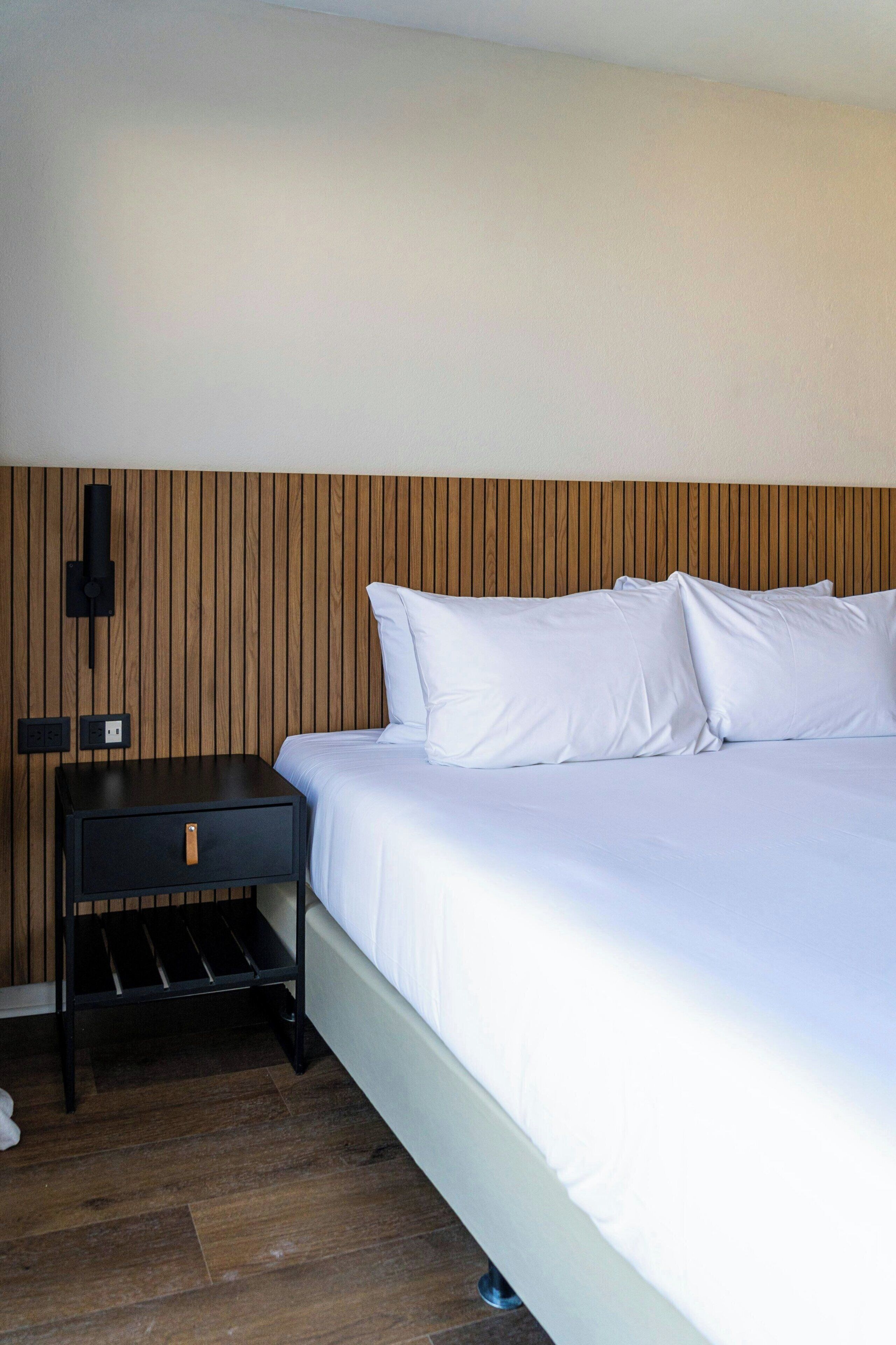 A hotel room with a bed, a nightstand, a wall-mounted lamp, and a wooden headboard.