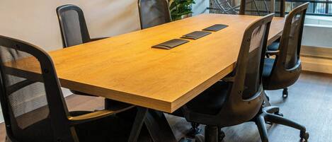 Un salón de conferencias con una mesa de madera, sillas de oficina negras, un televisor de pantalla plana en la pared y un amplio ventanal con vista a árboles.