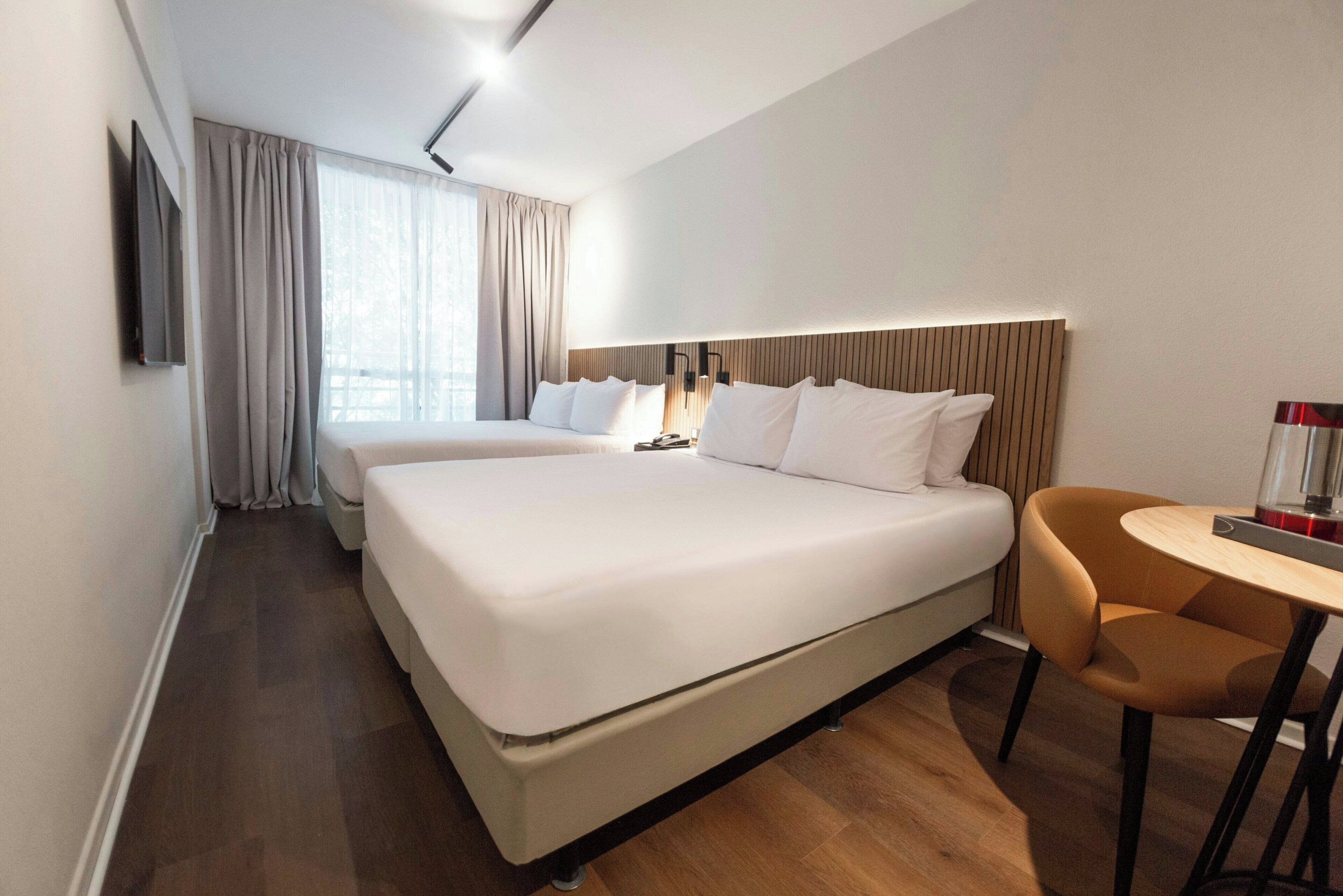 Ein Hotelzimmer mit einem großen Bett, einem Nachttisch, einem Sessel, einem Fernseher und einem Fenster mit Vorhängen.