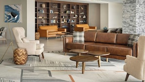 A modern living room with a brown leather sofa, beige armchairs, and a wooden herringbone side table. A large bookshelf filled with books and decorative items is in the background. - Four Points By Sheraton Oakville (Oakville)