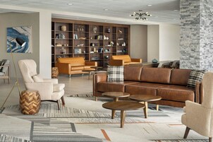 Una sala moderna con un sofá de cuero marrón, sillones beige y una mesita auxiliar de madera con patrón de espiga. En el fondo, una estantería grande llena de libros y objetos decorativos.