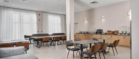 A modern hotel dining area with multiple tables and chairs, a kitchen in the background, and large windows with blinds.