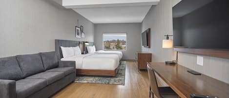 A hotel room with a sofa, two beds, a desk, a TV, and a window with a view.