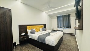 A modern hotel room with a large bed, a bedside table, a chair, a desk, and a window with curtains. - Vaayu Homestay (New Delhi)