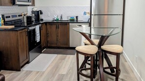 A modern kitchen with wooden cabinets, a stainless steel refrigerator, a microwave, and a glass-top dining table with two stools. - 2 Bedroom Guest Suite in SW (Calgary)