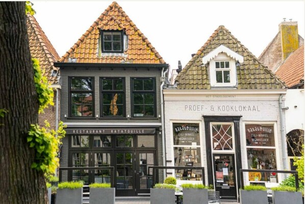 Een rij gebouwen met traditionele architectuur, waaronder een restaurant genaamd 'Ratatouille' en een winkel genaamd 'PROEF & KOOKLOKAAL'.