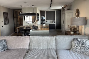 Een moderne woonkamer met een grijze bank, een eethoek met stoelen, een keuken met donkere kastjes, en een witte kast met decoratieve objecten.