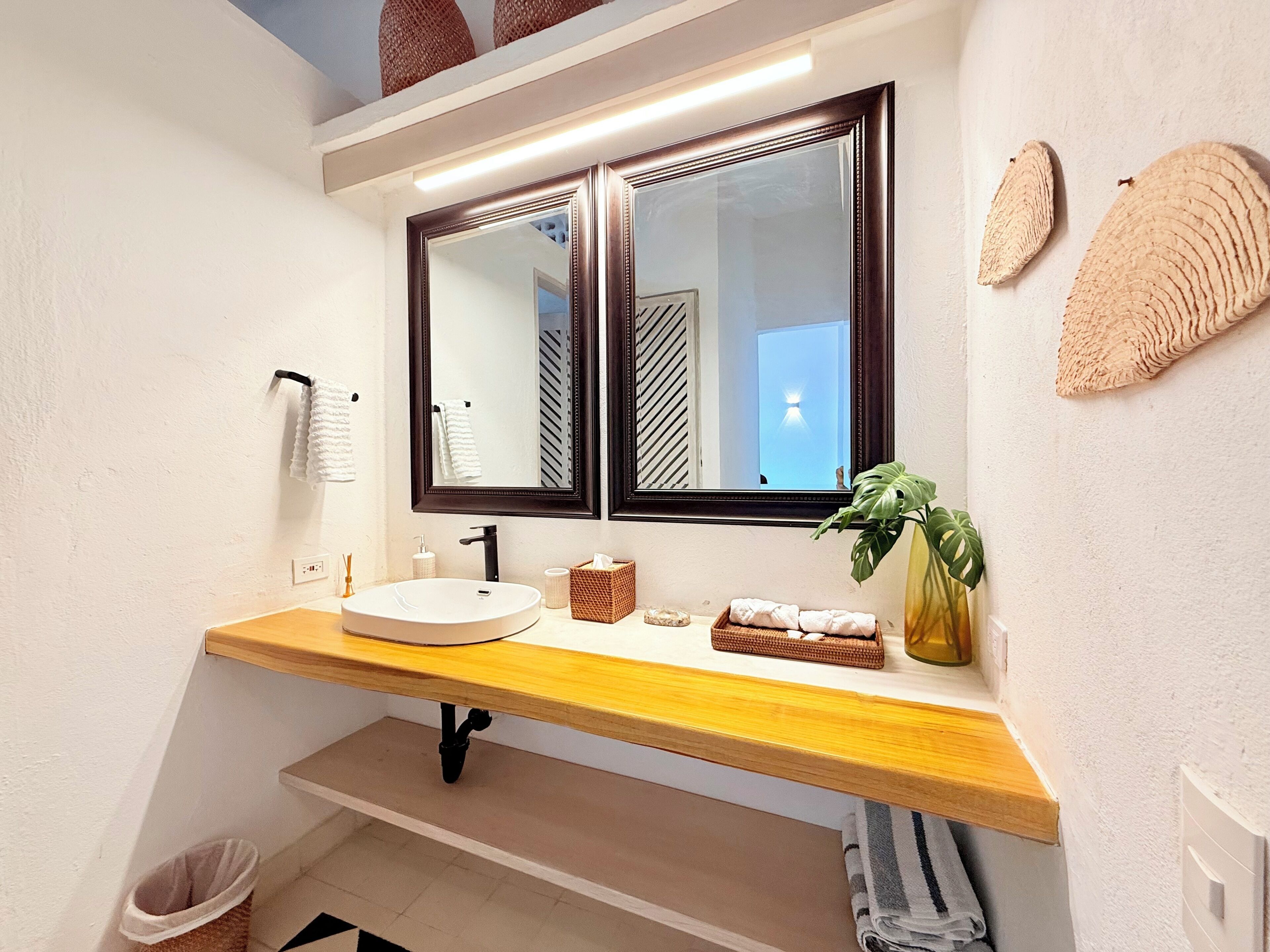 A bathroom with a wooden vanity, two mirrors, a sink, a towel rack, a waste bin, and a plant.