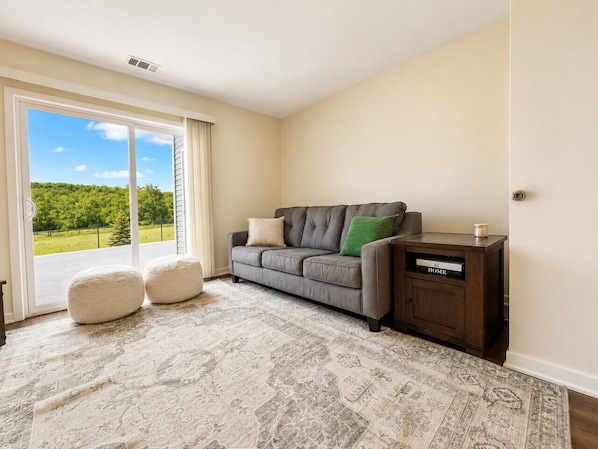 A room with a grey sofa, two ottomans, a patterned rug, a dark wooden side table, and a sliding glass door leading to a green landscape.
