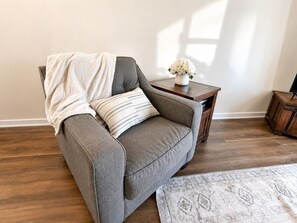 A gray sofa with a striped pillow, a small wooden side table with a vase of flowers, and a patterned rug on a wooden floor.