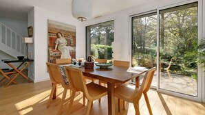 Un comedor con una mesa y sillas de madera, un cuadro en la pared y vistas al jardín a través de ventanas amplias.