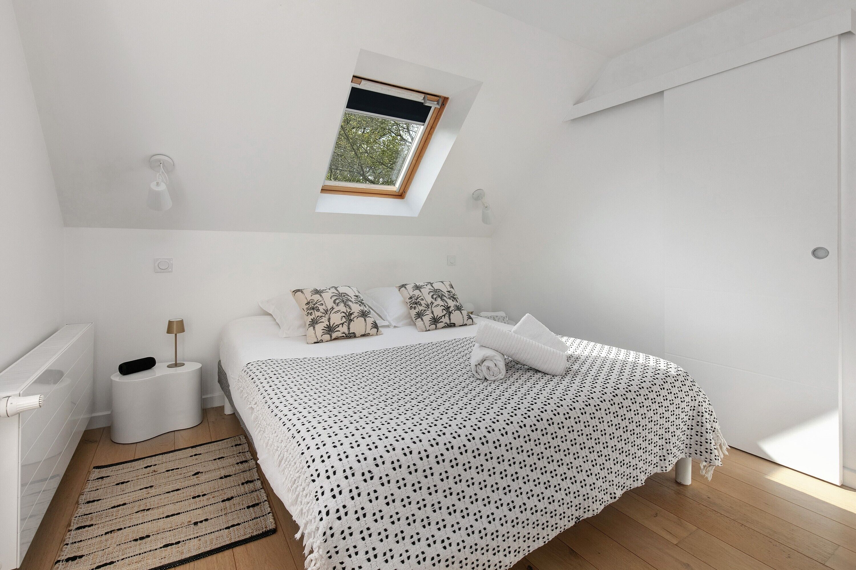 A bedroom with a skylight, a bed with patterned pillows, a bedside lamp, and a woven rug.