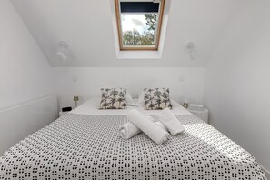 A small, white, single-bed room with a sloped ceiling, a skylight, two patterned pillows, a neatly folded towel, and a bedside table with a lamp. - Hakuna Matata: Peaceful Retreat with Large Terrace Overlooking the Forest (Saint-Molf)