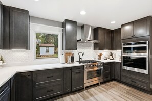 A modern kitchen with dark cabinetry, a built-in oven, a stove, and a window with a view of a small house. - Spacious Colorado Retreat, Game Room, Solarium, Patios & Mountain View’s (Evergreen)