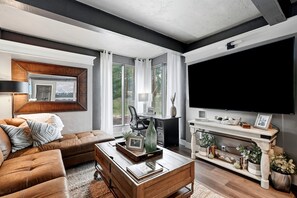 A modern living room with a leather sofa, a wooden coffee table, a flat-screen TV, and a large window offering a view of trees. - Spacious Colorado Retreat, Game Room, Solarium, Patios & Mountain View’s (Evergreen)