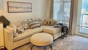 Un salon avec un canapé beige, des coussins à motifs, un pouf rond, une table basse et une grande fenêtre avec des stores.