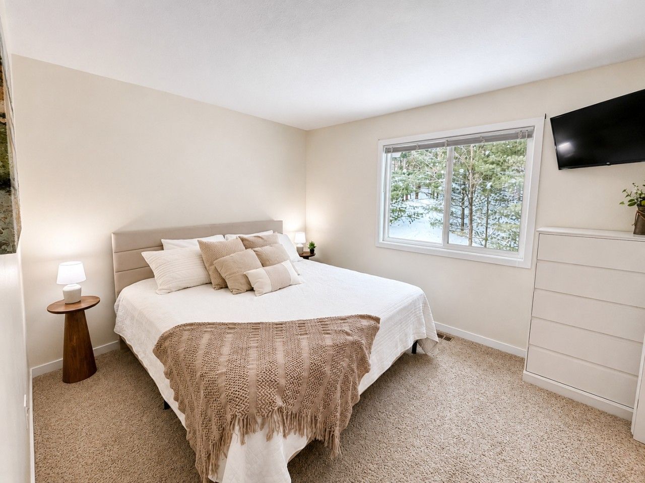 A bedroom with a bed, bedside lamp, window, and a TV mounted on the wall.