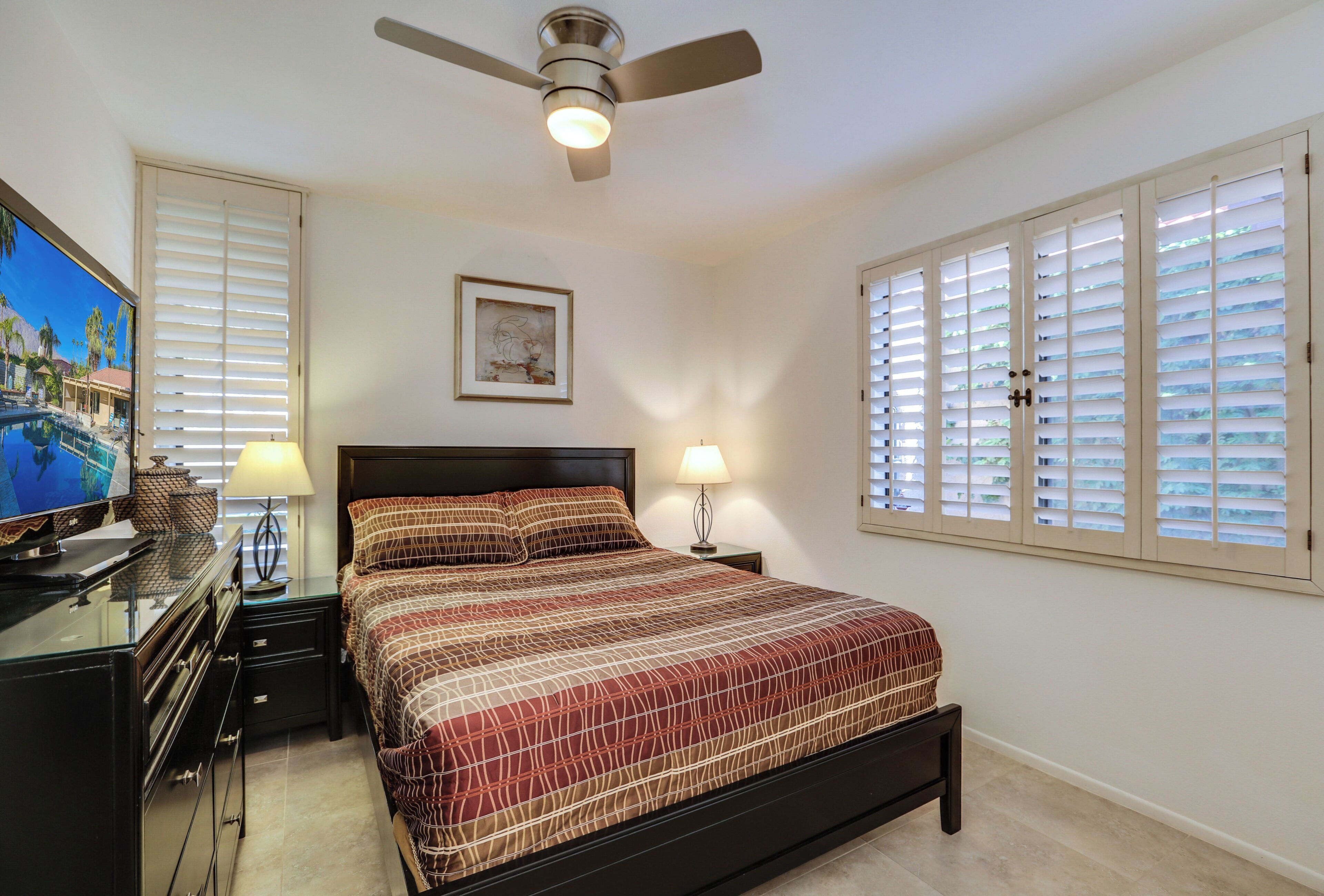 A bedroom with a bed, a ceiling fan, a desk with a lamp, a TV, and a window with blinds.