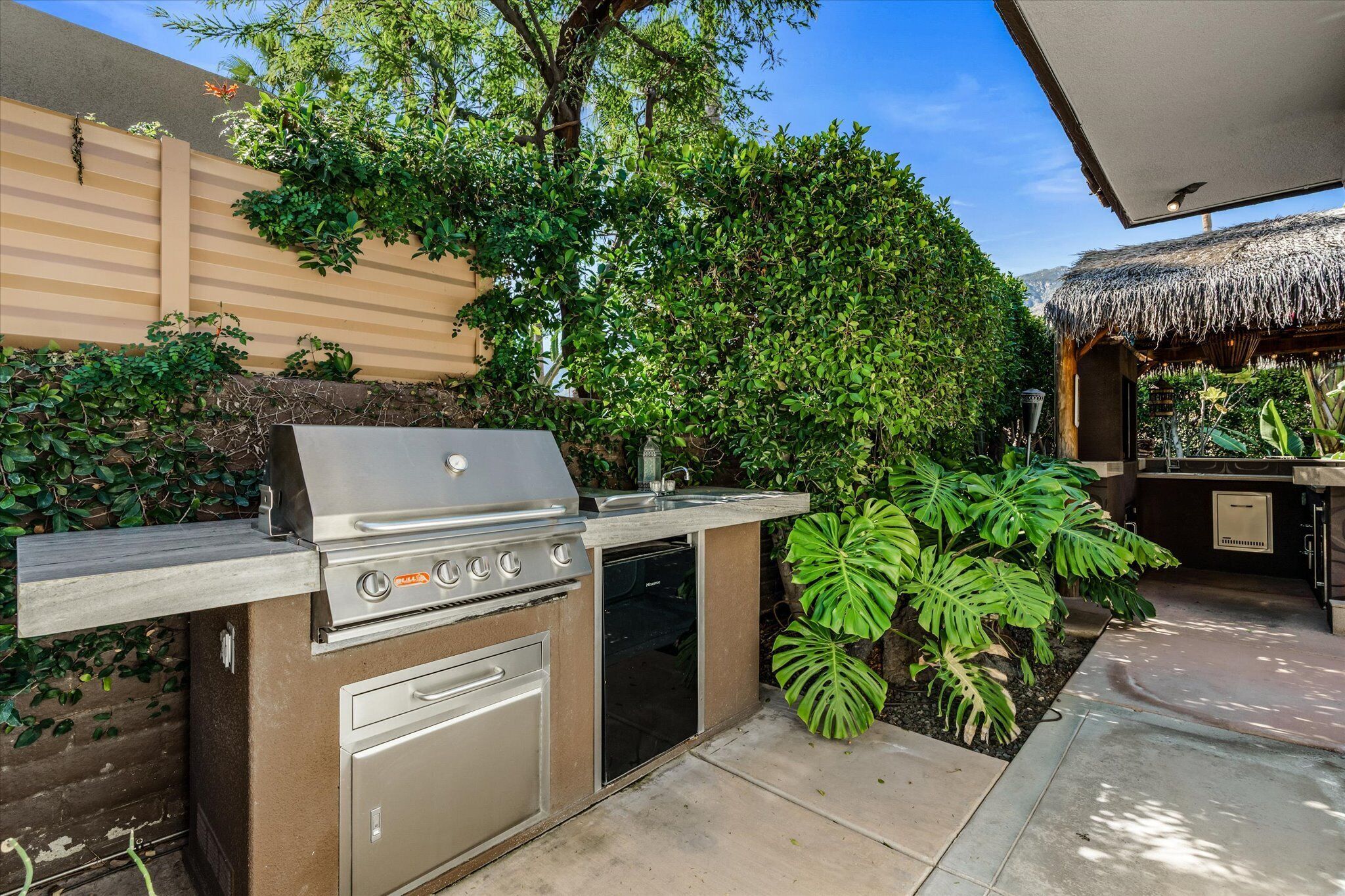 Cocina exterior con parrilla integrada, pileta y un mueble de almacenamiento, rodeada de vegetación exuberante y una estructura de techo de paja.