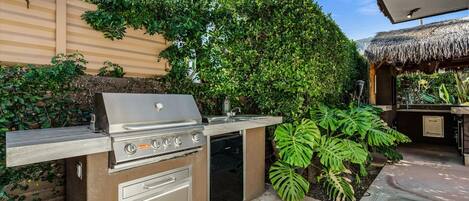 Cocina exterior con parrilla integrada, pileta y un mueble de almacenamiento, rodeada de vegetación exuberante y una estructura de techo de paja.