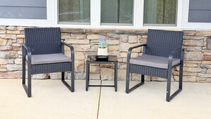 Two outdoor wicker chairs with cushions, a small table, and a potted plant, set against a stone wall and a window with curtains. - Elegant 5BR/4BA Masters Rental – Just 7 Miles from Augusta National (Martinez)