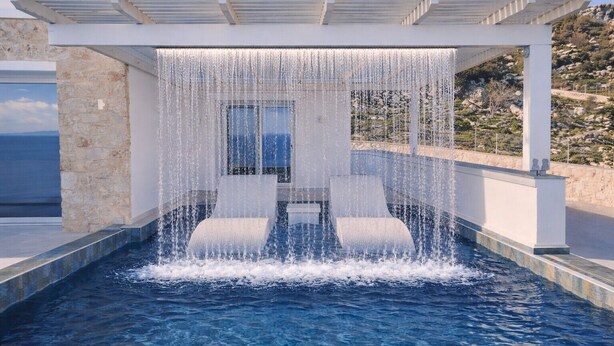 Una piscina con agua que cae sobre una estructura, un muro de piedra y vista al mar.