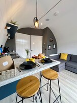 A modern kitchen with a marble countertop, two stools, a dining table, and a sofa with a yellow pillow.