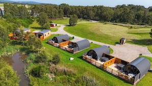 A series of dome-shaped cabins with outdoor seating, surrounded by a grassy field and a small body of water. - Newmore Highland Pods (Invergordon)
