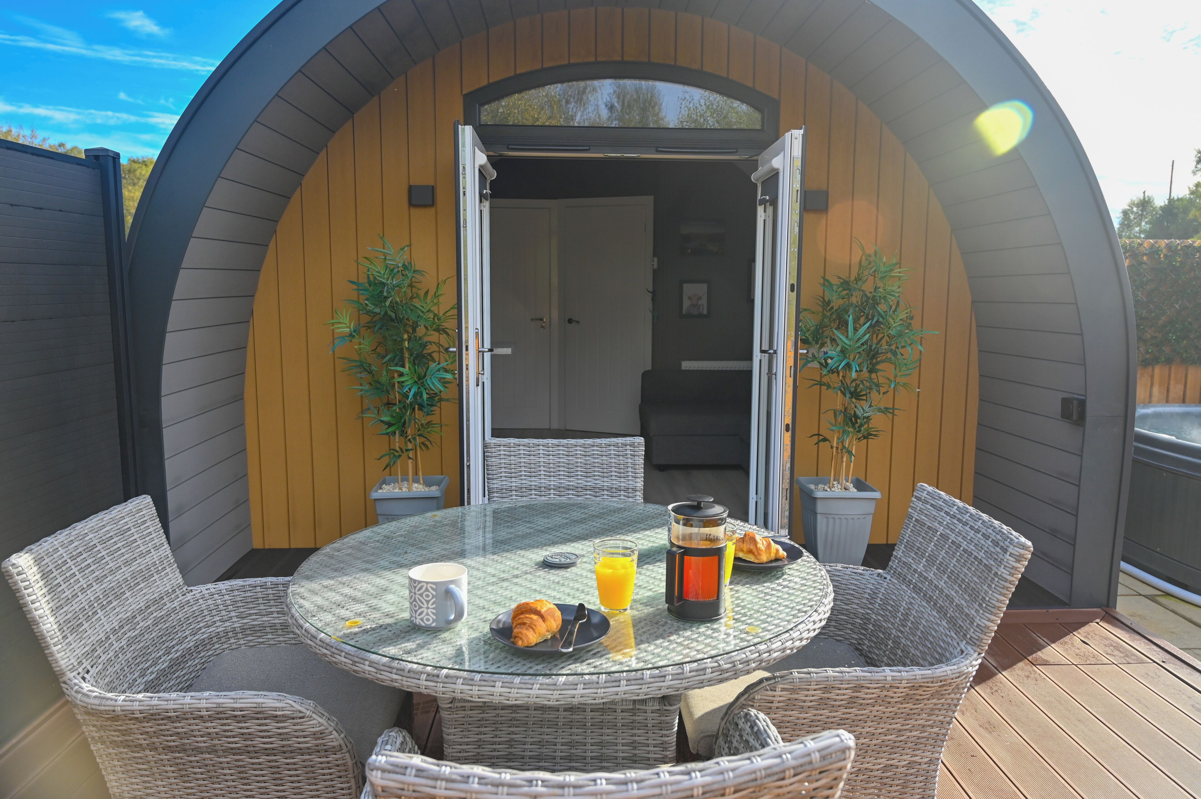 A circular outdoor dining area with wicker furniture, a glass table, and a breakfast setting including croissants, juice, and a coffee pot.