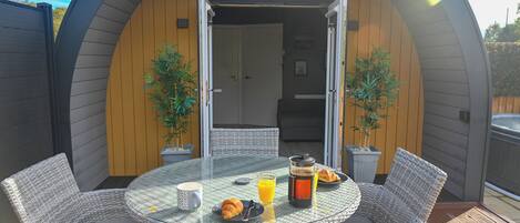 A circular outdoor dining area with wicker furniture, a glass table, and a breakfast setting including croissants, juice, and a coffee pot.
