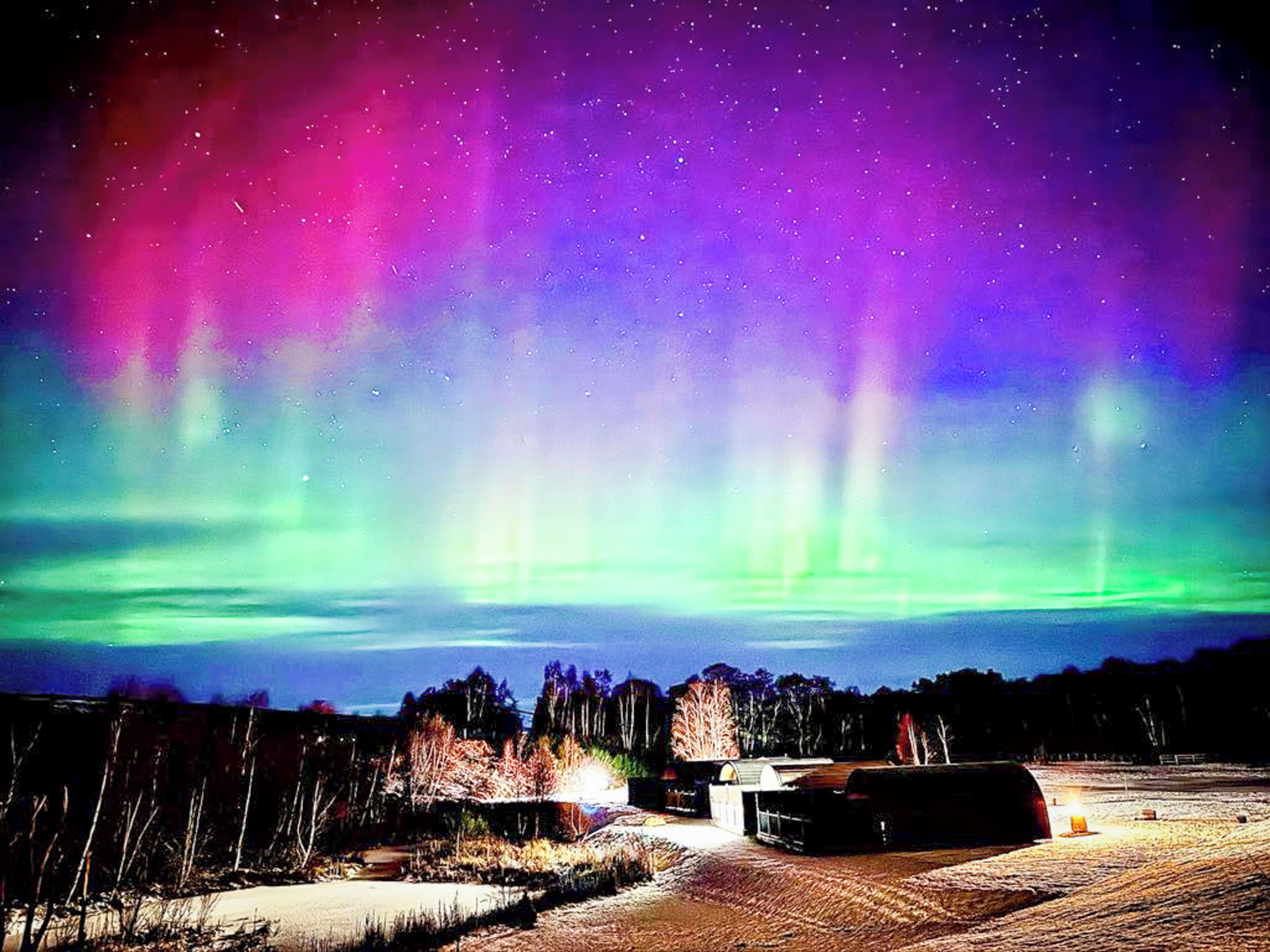 A snowy landscape with a building and trees under a colorful aurora display.