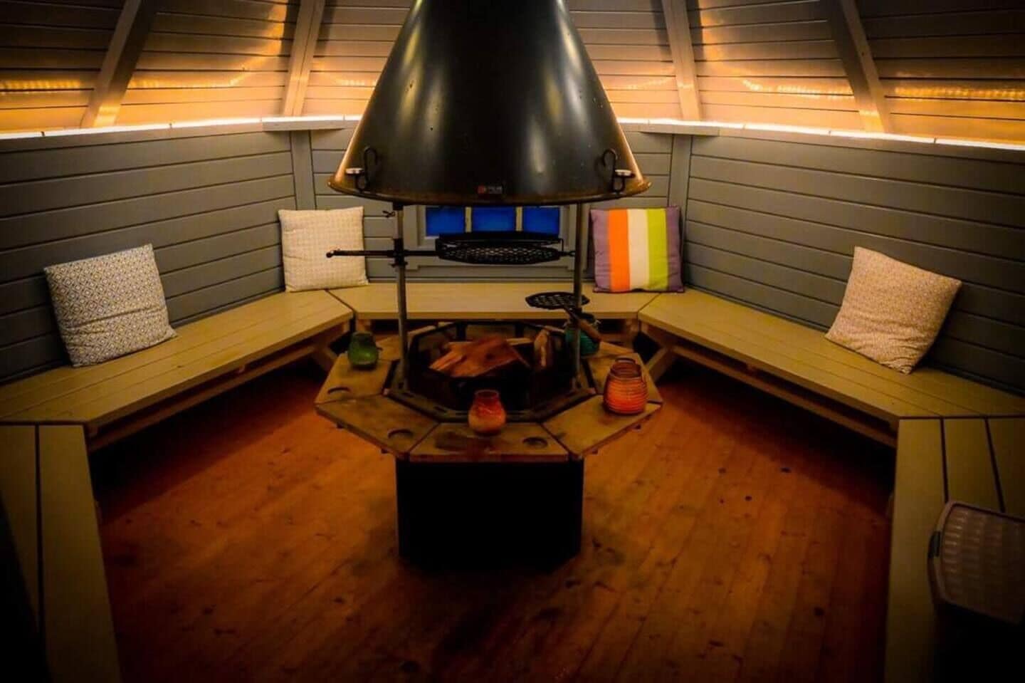 A cozy room with a central wood-burning stove, benches, and a sloped ceiling with lighting.