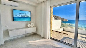 Een modern interieur met een televisie waarop 'Welcome to AQUA BLUE VILLA' staat, een balkon met uitzicht op zee, en een minimalistisch ontwerp.