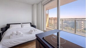 Un dormitorio moderno con una cama grande, ropa de cama blanca, un televisor sobre un soporte de madera y un balcón con vista a la ciudad.