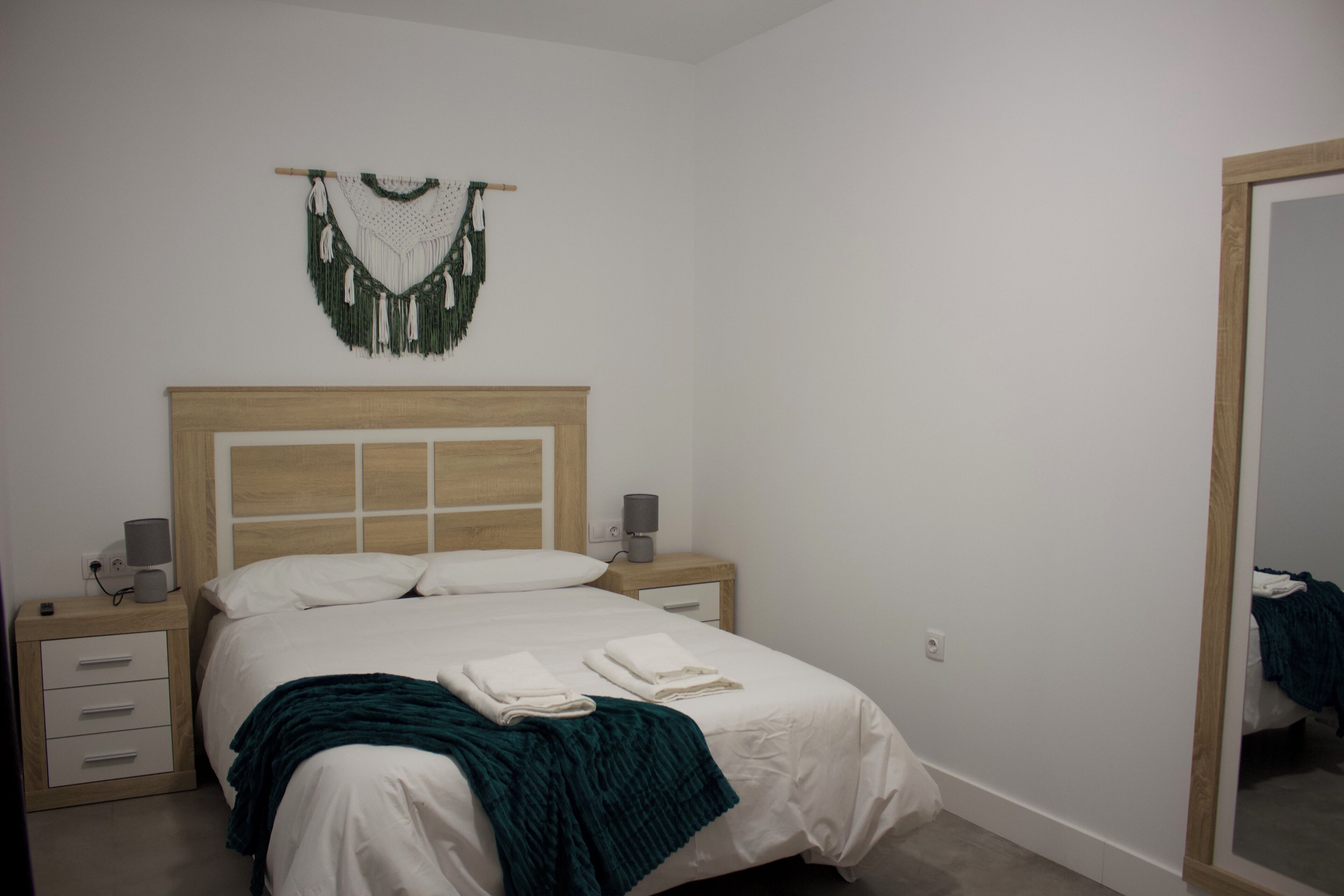 Un dormitorio con cabecera de madera, cama con ropa blanca, dos mesitas de noche con lámparas y un tapiz de macramé en la pared.