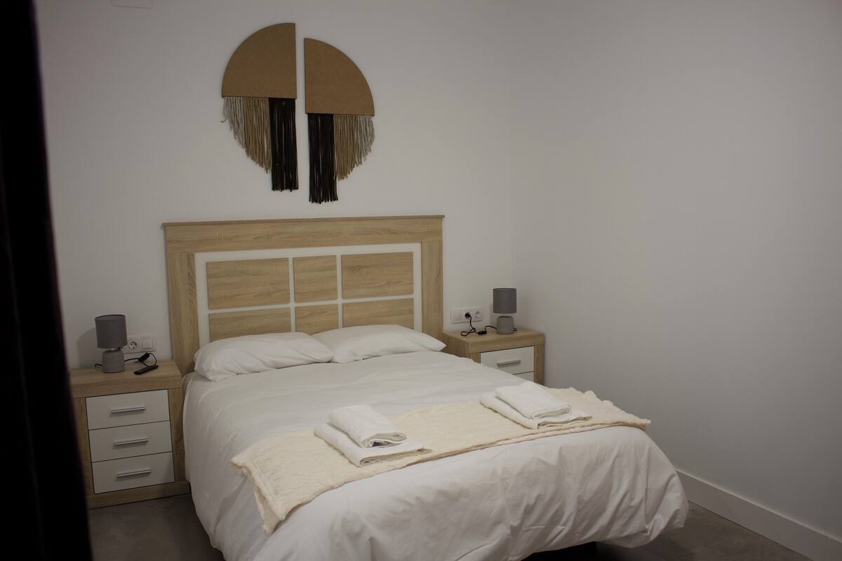 Un lit bien fait, avec du linge de lit blanc, une table de chevet avec une lampe et un élément décoratif fixé au mur.