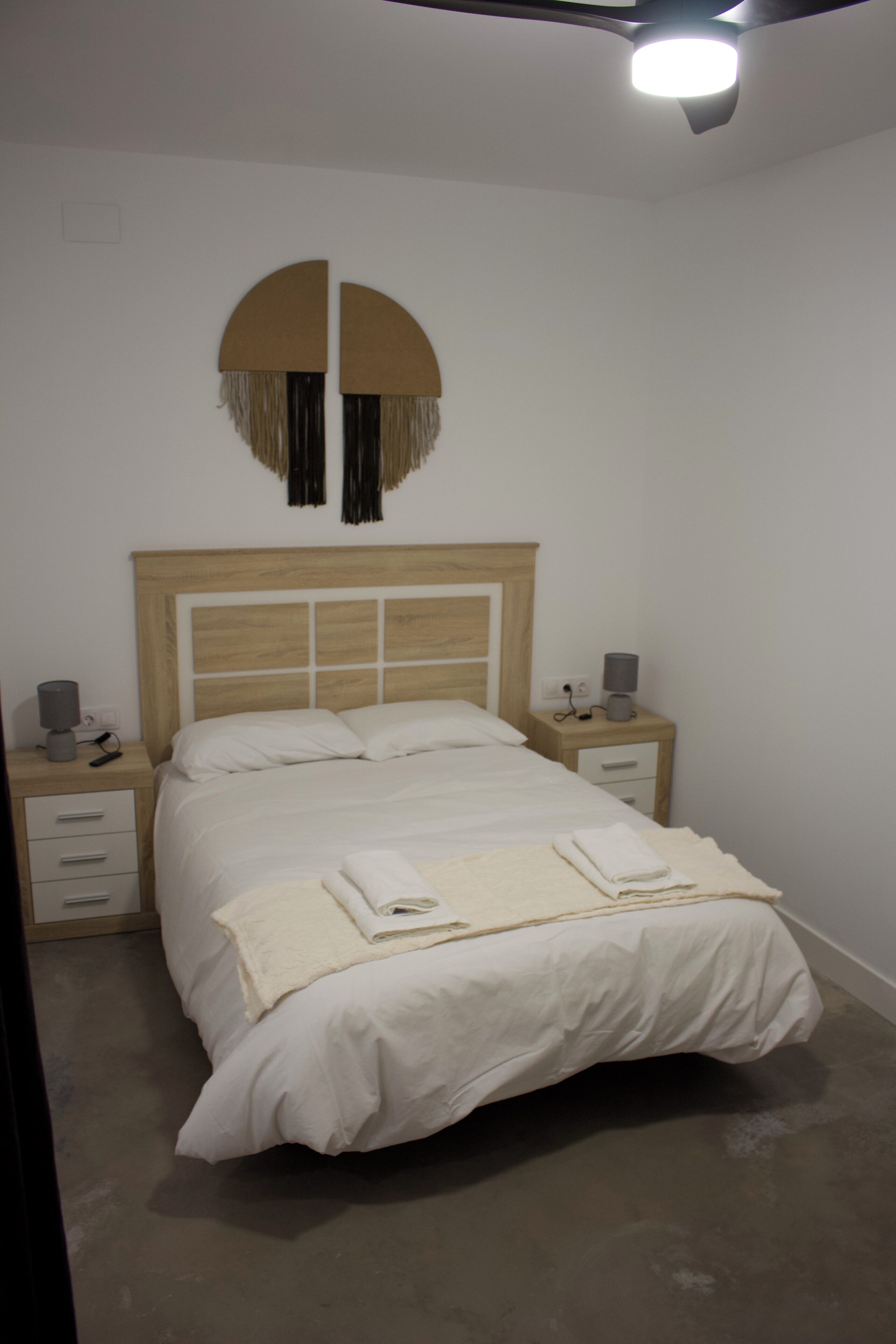 Un dormitorio con una cama, mesitas de noche, un adorno circular en la pared y un ventilador de techo.