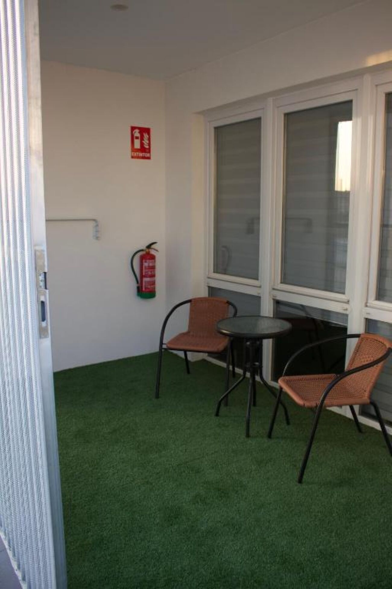 Un pequeño espacio exterior con césped artificial, dos sillas de mimbre, una mesita redonda y un extintor de incendios montado en la pared.