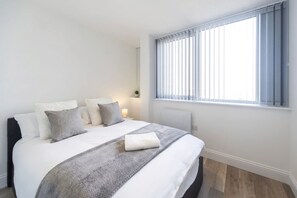 A bedroom with a bed, pillows, a bedside table with a lamp, a window with blinds, and a radiator. - Modern 2-Bed Gravesend Apt with Games Room (Gravesend)