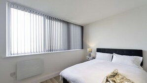 A hotel room with a large window, a bed with white linens, a bedside table with a lamp, and a radiator. - Modern 2 Bed Apartment in Gravesend w Games Room (Gravesend)
