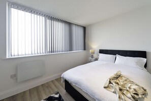 A hotel room with a large window, a bed with white linens, a bedside table with a lamp, and a radiator. - Modern 2 Bed Apartment in Gravesend w Games Room (Gravesend)