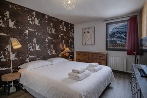 Ein Schlafzimmer mit einem gro ßen Bett, einer Holzkommode, einem Nachttisch mit Lampe, einer Wand mit Kuhfellmuster, einem gerahmten Bild und einem Fenster mit Bergblick.