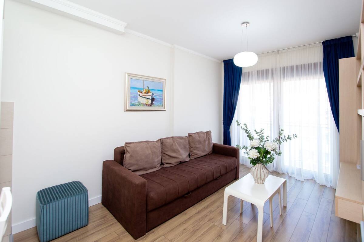 Un salón con un sofá marrón, una mesa blanca, un reposapiés azul y un cuadro de un barco en la pared.