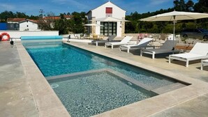 A swimming pool with lounge chairs and umbrellas, a building with a red roof, and a clear blue sky. - Casaspensadas (Porto de Mos)