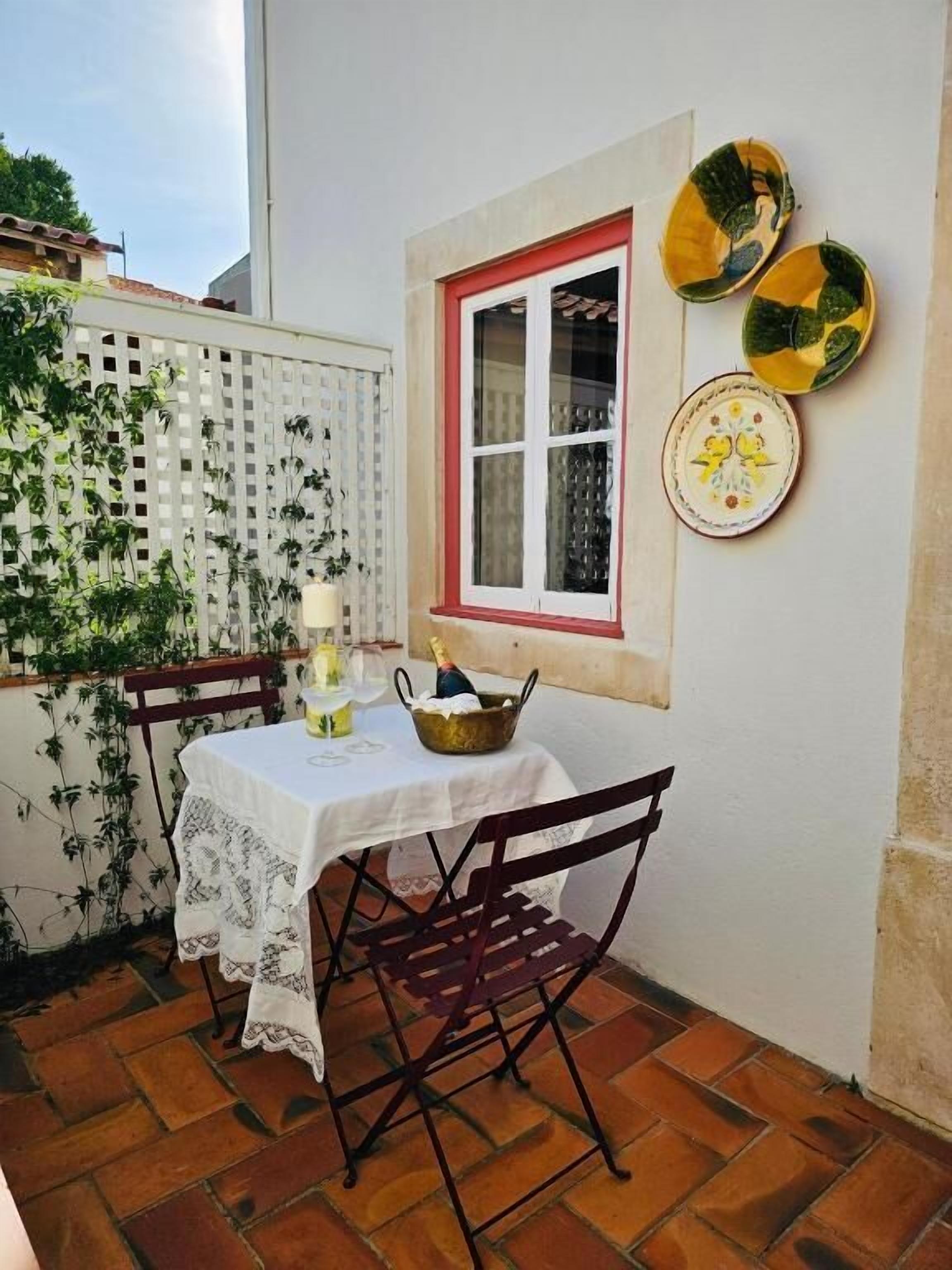 A small patio with a table set for two, a window with a red frame, and decorative plates on the wall.